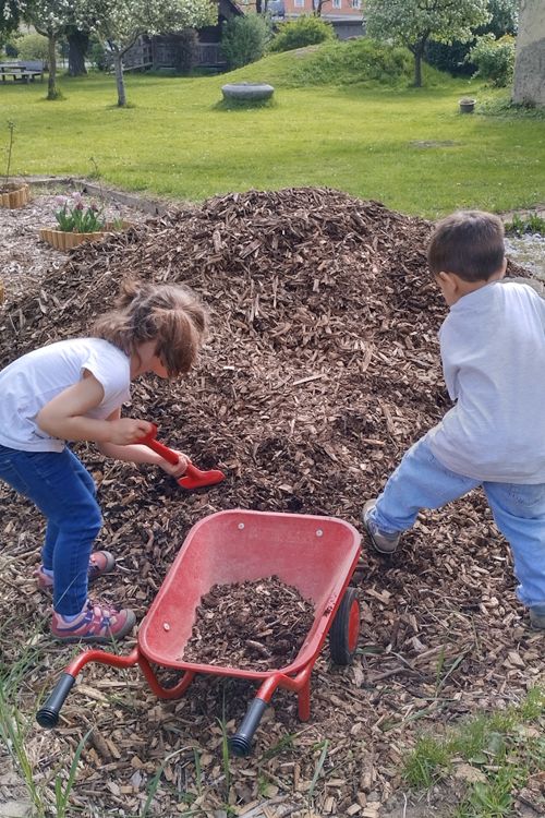 Zwei Kinder schaufeln Rindenmulch in eine Scheibtruhe